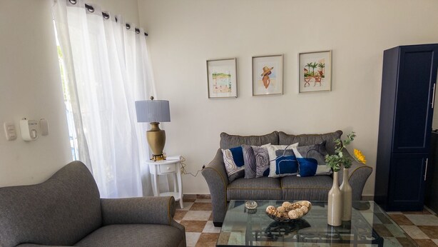 Un salon avec un canapé gris, une table basse en verre et un tapis à motifs. Il y a trois tableaux encadrés au mur, une lampe sur une table d’appoint et un meuble sombre.