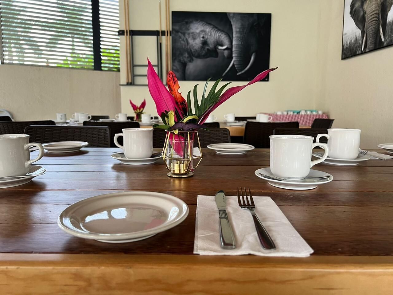 Una mesa de comedor dispuesta para cuatro personas, con platos, tazas y cubiertos blancos, un florero con flores y una foto enmarcada de un elefante en la pared.