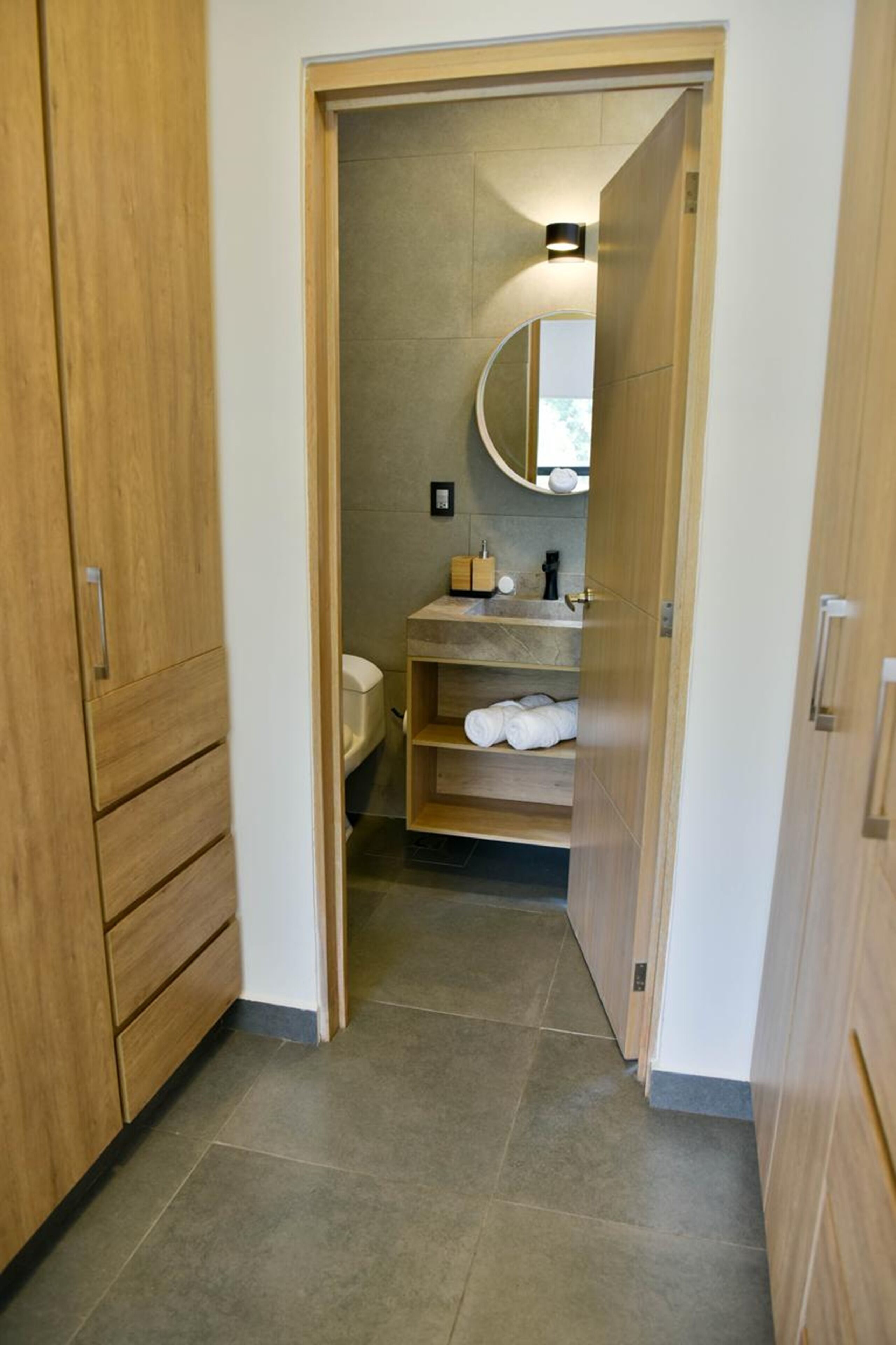 A narrow hallway leading to a bathroom with a toilet, sink, and shelves with towels.