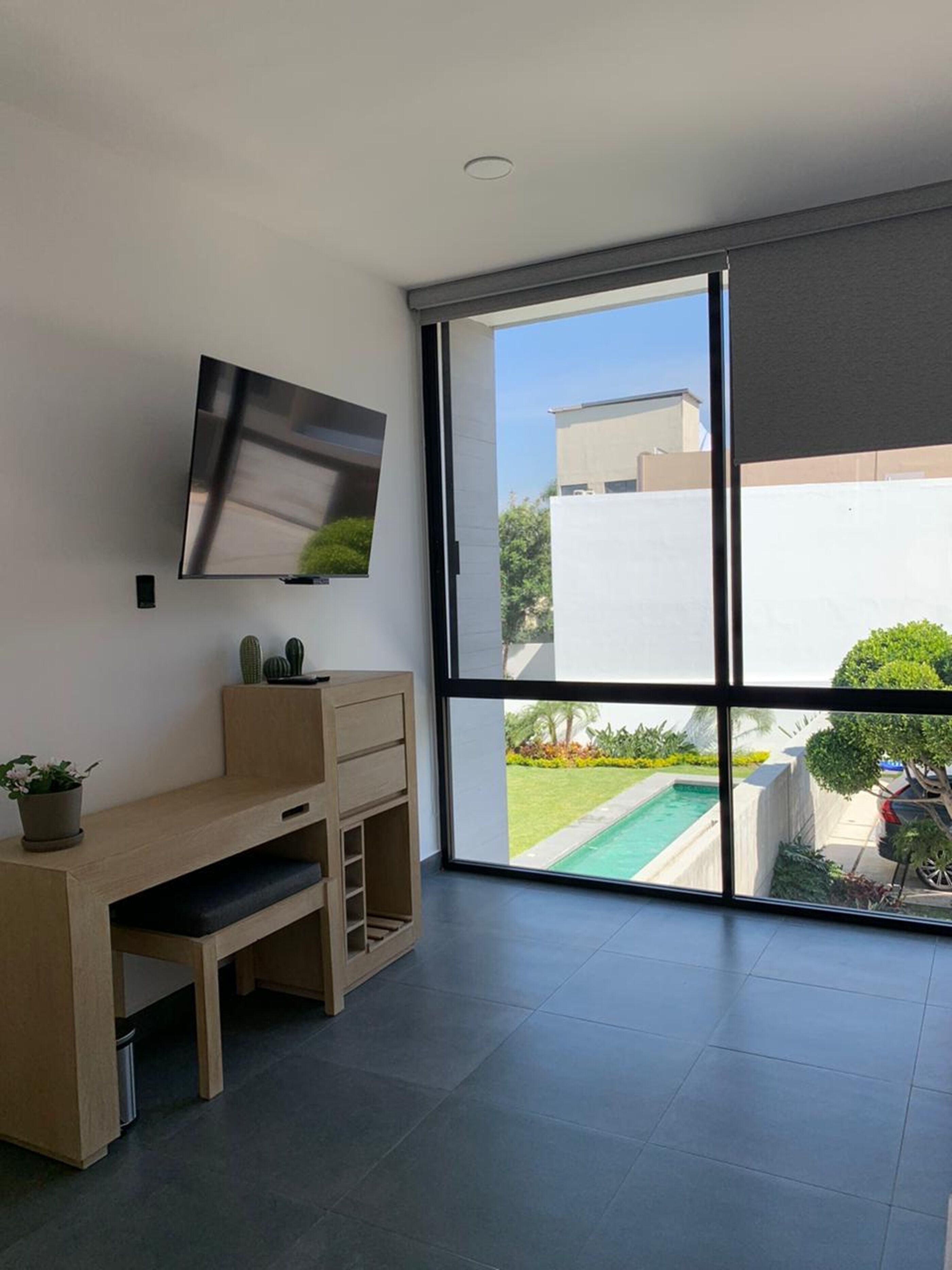 Una habitación moderna con un ventanal grande, un televisor de pantalla plana montado en la pared, un escritorio de madera y una planta en maceta.
