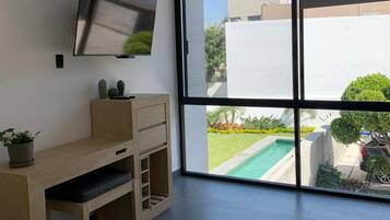 Una habitación moderna con un ventanal grande, un televisor de pantalla plana montado en la pared, un escritorio de madera y una planta en maceta.