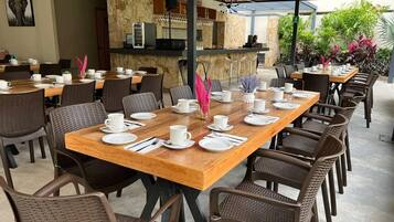 Un restaurante con varias mesas dispuestas para comer, una zona de bar y terraza con plantas.