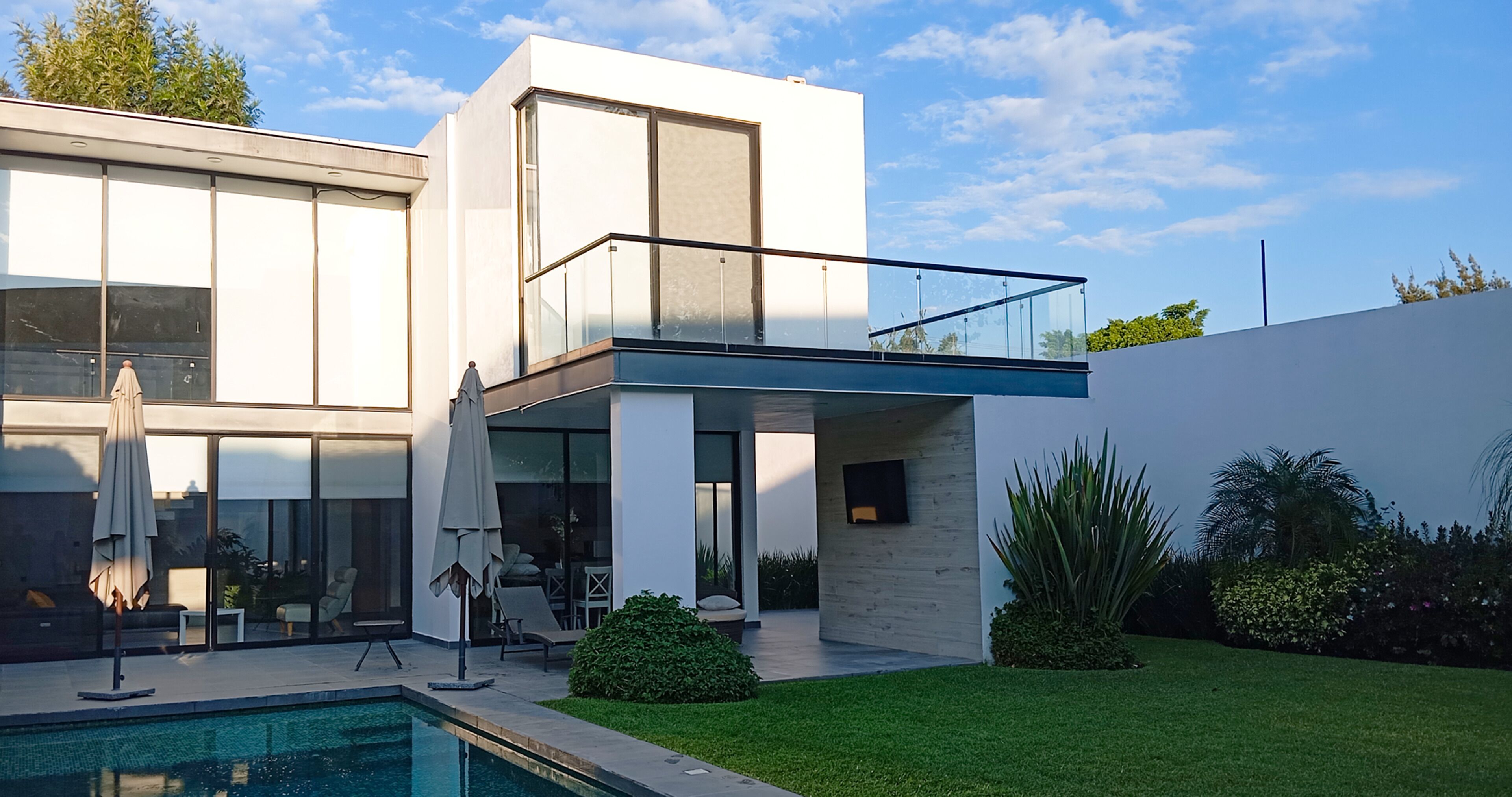 Et moderne hus med to etager, en swimmingpool, store vinduer og en balkon.