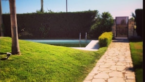 Un sendero de piedra conduce a una zona de piscina, con vista despejada al agua y un seto al fondo.