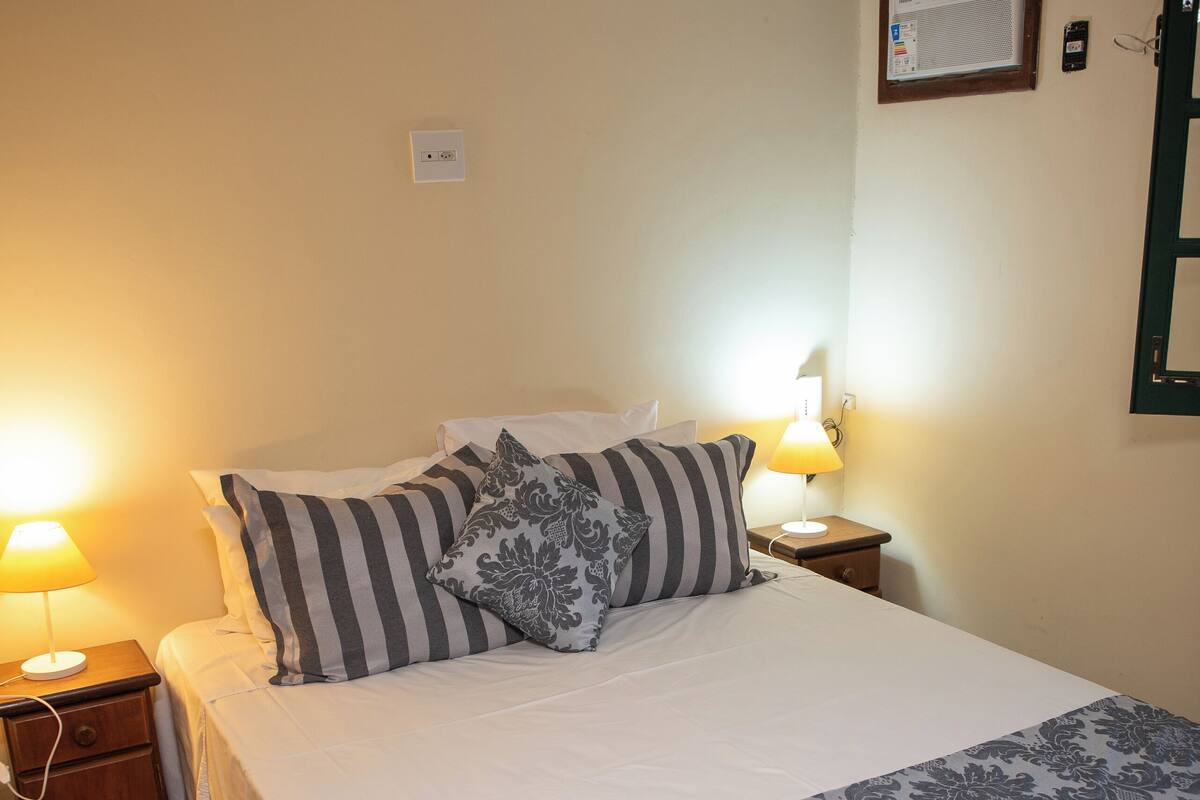 A neatly made bed with striped and floral pillows, a bedside lamp, and a nightstand with a power outlet.
