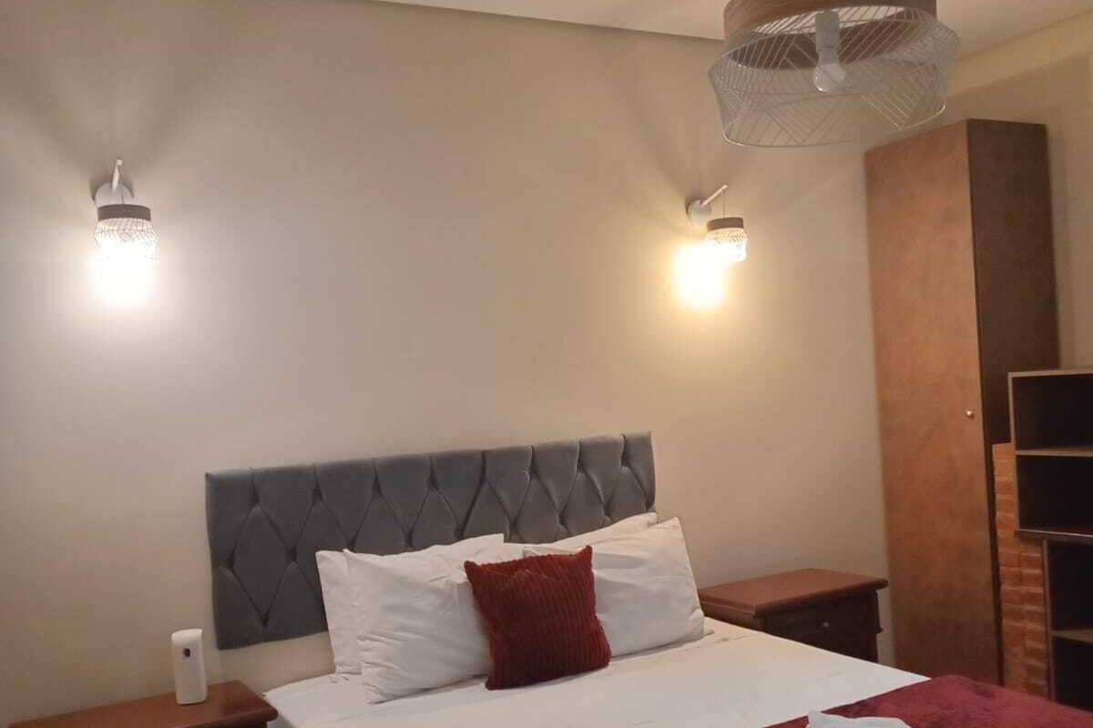 A hotel room with a bed, two bedside tables, a nightstand, a red throw blanket, and a pair of rolled towels.