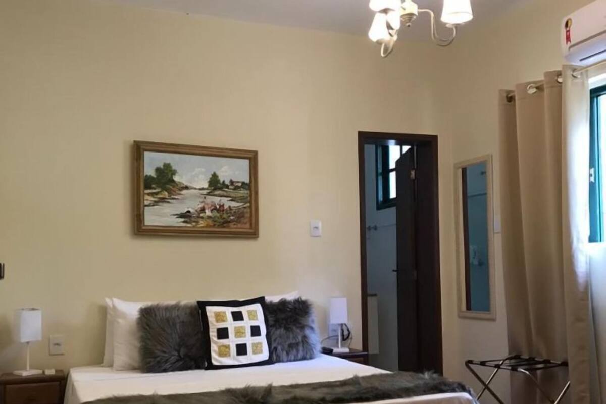 A bedroom with a bed, a nightstand, a lamp, a painting on the wall, and a window with curtains.