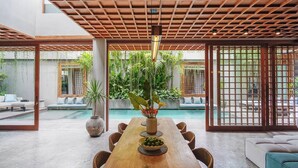 A spacious indoor area with a wooden dining table, chairs, and a large window overlooking a swimming pool. - Designers Dream Near Beach (Bali)