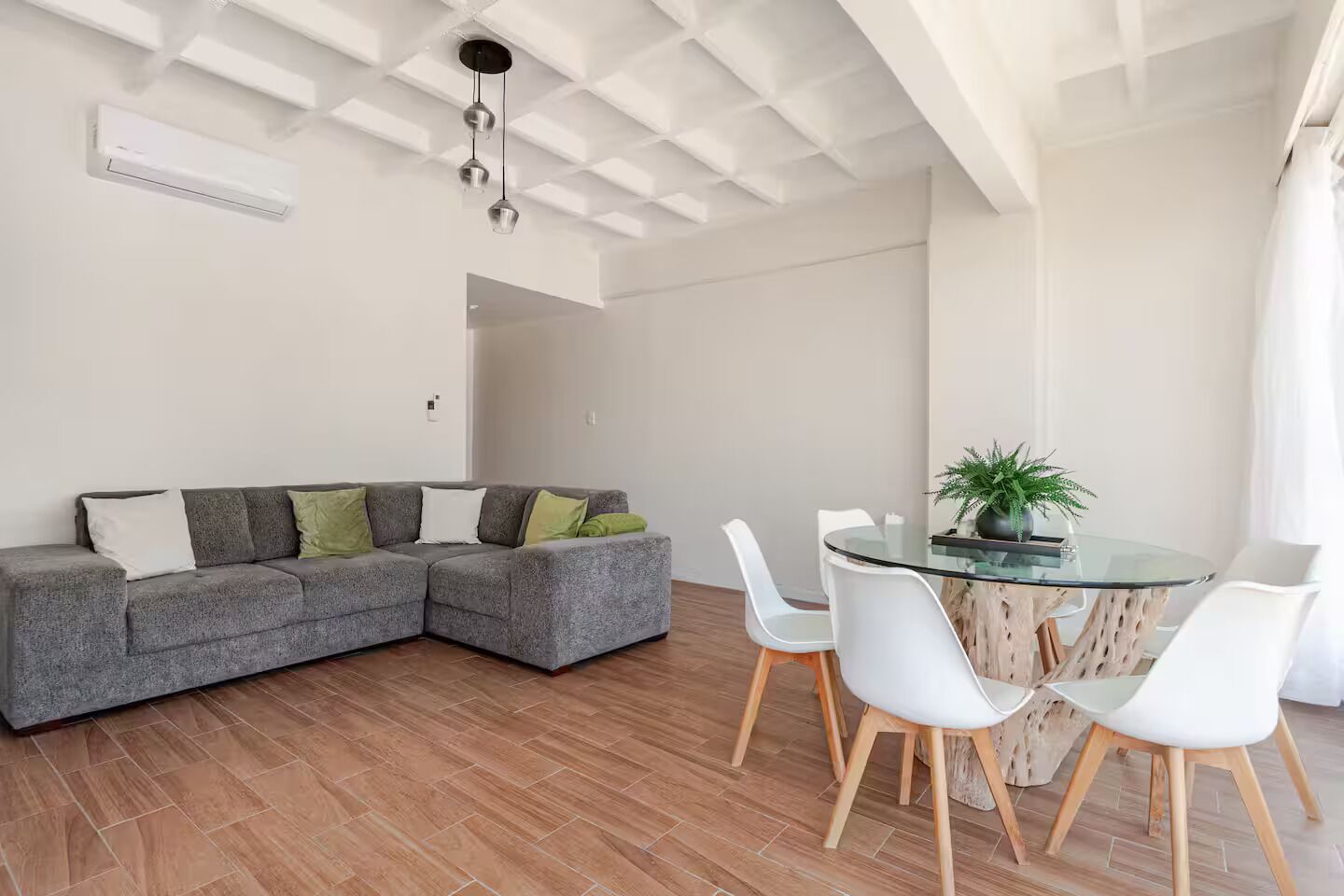Ein modernes Wohnzimmer mit einem grauen Ecksofa, einem Esstisch mit Glasplatte und weißen Stühlen sowie einer Topfpflanze.