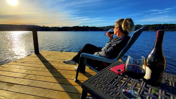 A person sitting on a wooden deck by a lake with a bottle of wine and a glass.