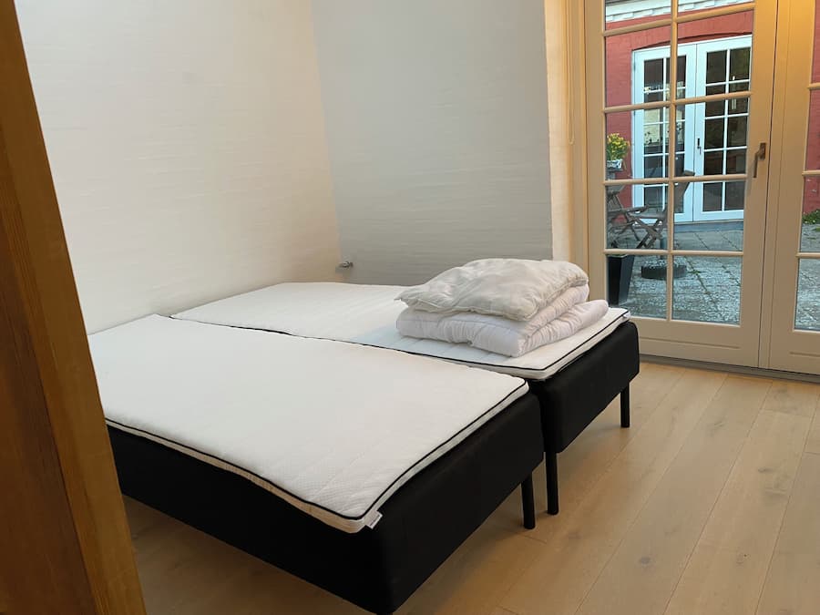 Cama doble con colchón blanco y estructura negra, mesita de noche pequeña y vista al exterior a través de una puerta de cristal.