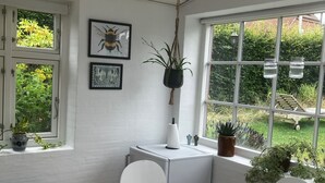 A dining area with a round wooden table, white chairs, a black side table, and a hanging light fixture. There are two windows with views of greenery and a white wall with two framed pictures. - Husmandsvej 5 (Odder)