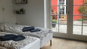 A room with two beds, a window, and a door leading to an outdoor area with a red wall and plants. - Husmandsvej 5 (Odder)