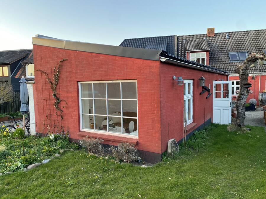 Una casa in mattoni rossi con un'ampia finestra, una porta bianca e un piccolo giardino con piante e una bicicletta.