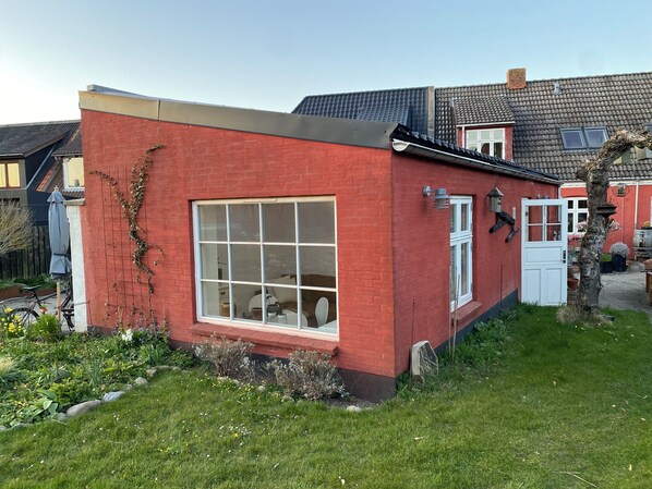A red brick house with a large window, a white door, and a small garden area with plants and a bicycle. - Husmandsvej 5 (Odder)