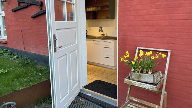 A red exterior wall with a white door, a small garden area, and a wooden planter with yellow flowers. - Husmandsvej 5 (Odder)
