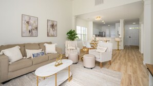 A spacious living room with a beige sofa, a white coffee table, and two armchairs. There are abstract paintings on the wall, a ceiling fan, and a view into the kitchen area. - Bluffton Lowcountry Casa Del Café/Private Driveway+Fence Yard (Bluffton)