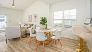 A modern living room with a round dining table, white chairs, a sofa, and a large window with blinds. - Bluffton Lowcountry Casa Del Café/Private Driveway+Fence Yard (Bluffton)