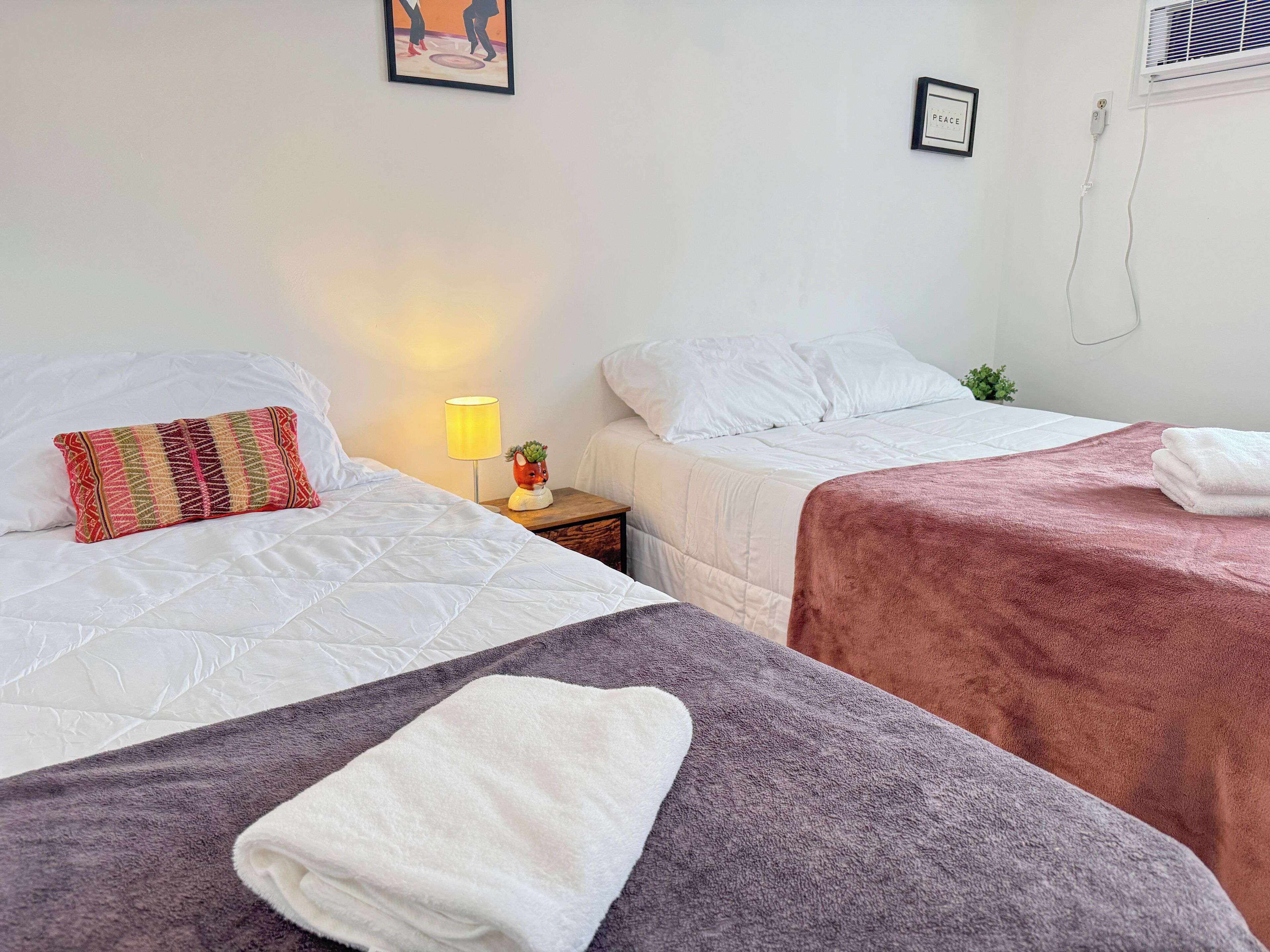 Habitación de hotel con dos camas, una mesita de noche con lámpara y aire acondicionado instalado en la pared.