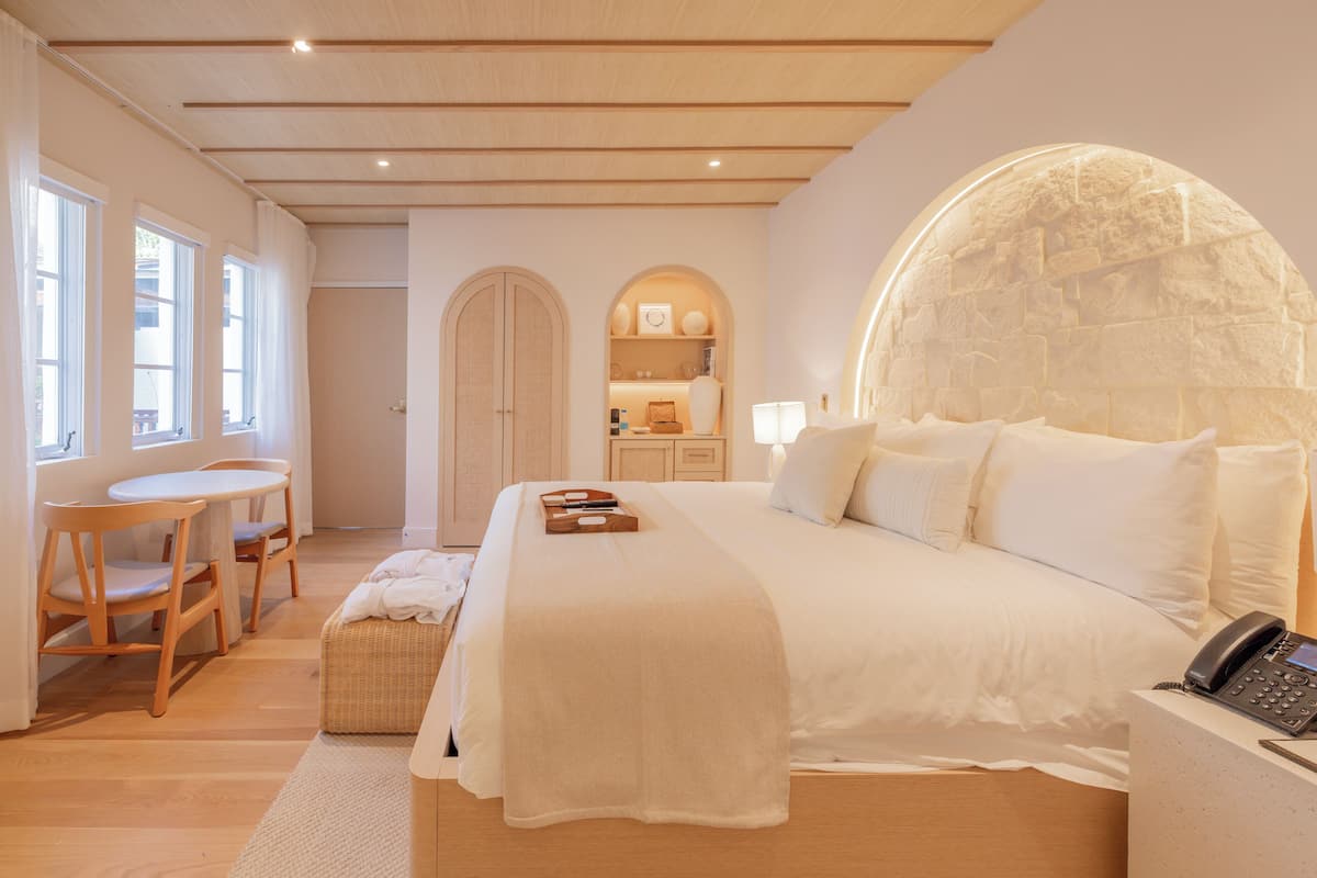 A bedroom with a large bed, a bedside table with a phone, a desk with chairs, and a small kitchenette in the background.