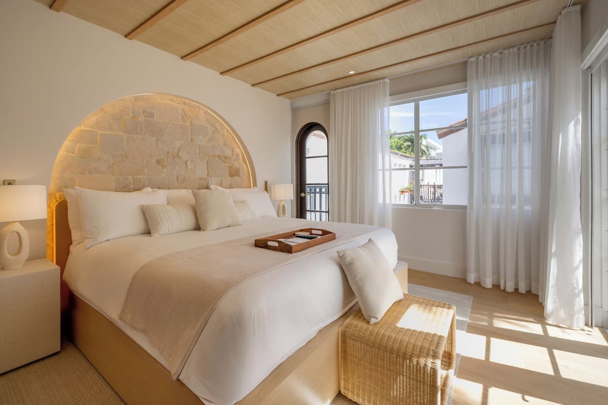 A large bed with a stone headboard, a bedside table with a lamp, and a wicker bench.
