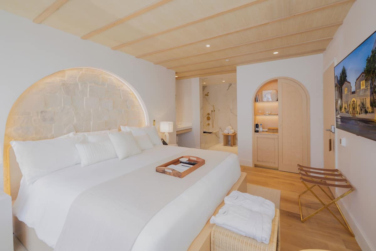 A modern hotel room with a large bed, a wooden headboard, a small seating area, and a view of the outside.
