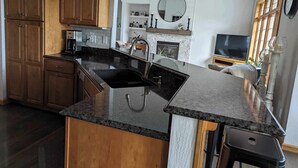 A kitchen with wooden cabinets, a granite countertop, a built-in sink, a coffee maker, a fireplace, a television, and a clock on the wall.