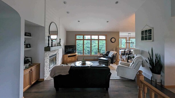 A spacious living room with a fireplace, a large window offering a view of trees, a dining area, and a ceiling fan.