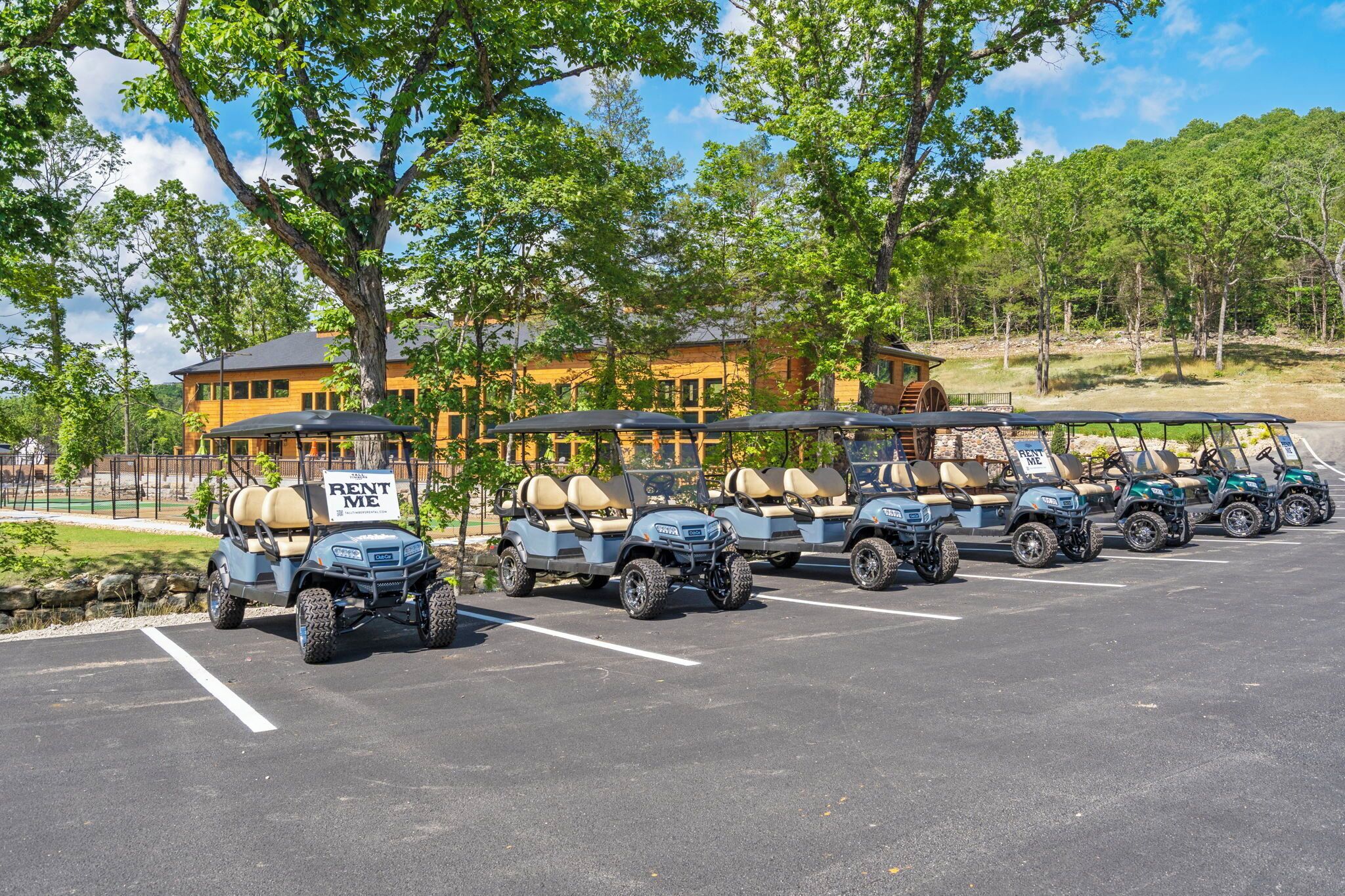 A row of golf carts parked in a lot with a 'RENT ME' sign, surrounded by trees and buildings.