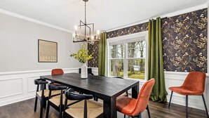 Een eetkamer met een donker houten tafel, zes stoelen, een kroonluchter en een raam met groene gordijnen.
