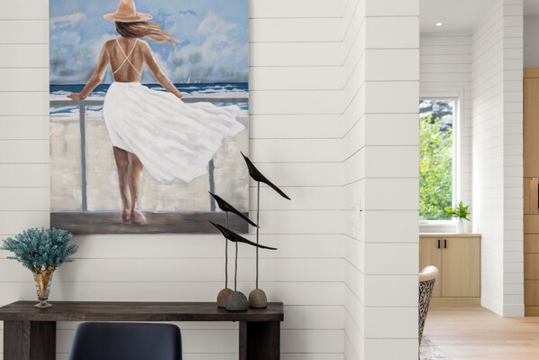 A modern interior with a large painting of a woman by the sea, a wooden table with decorative items, and a view of the outdoors through a window.
