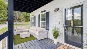 Eine überdachte Veranda mit einem weißen Sofa, einem kleinen Tisch, einem Blumentopf und einer Willkommensmatte.