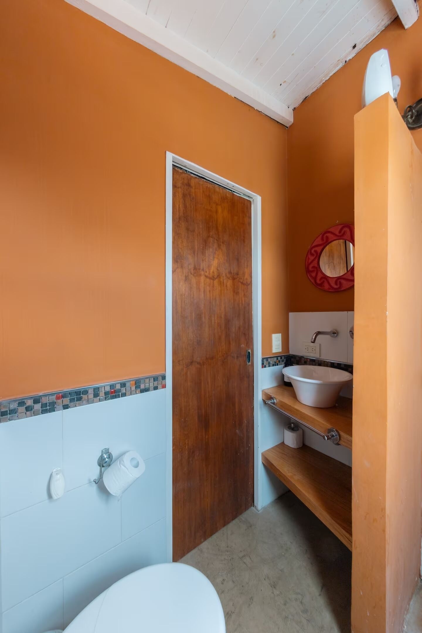 Un baño con una pared naranja, una puerta de madera, un lavabo blanco y un inodoro.