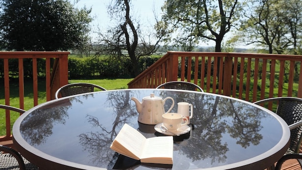 A round glass table with a book, a teapot, and a mug on a deck with chairs.