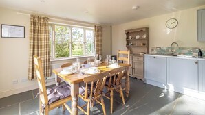 A dining area with a wooden table set for four, a window with curtains, a clock on the wall, and a kitchen area with cabinets and a sink.