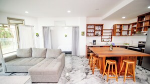 Una sala de estar moderna con un sofá, una mesa de comedor de madera con taburetes y una cocina con estantes y electrodomésticos.