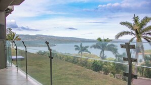 Un balcón con baranda de cristal que da a un lago, palmeras y colinas distantes.