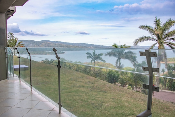 A balcony with a glass railing overlooking a lake, palm trees, and distant hills.