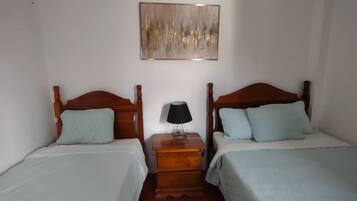Una habitación con dos camas de madera, cada una con una almohada azul claro y una colcha blanca. Entre las camas, hay una mesita de noche de madera con una lámpara. Encima de la mesita de noche, hay un cuadro abstracto enmarcado en la pared.