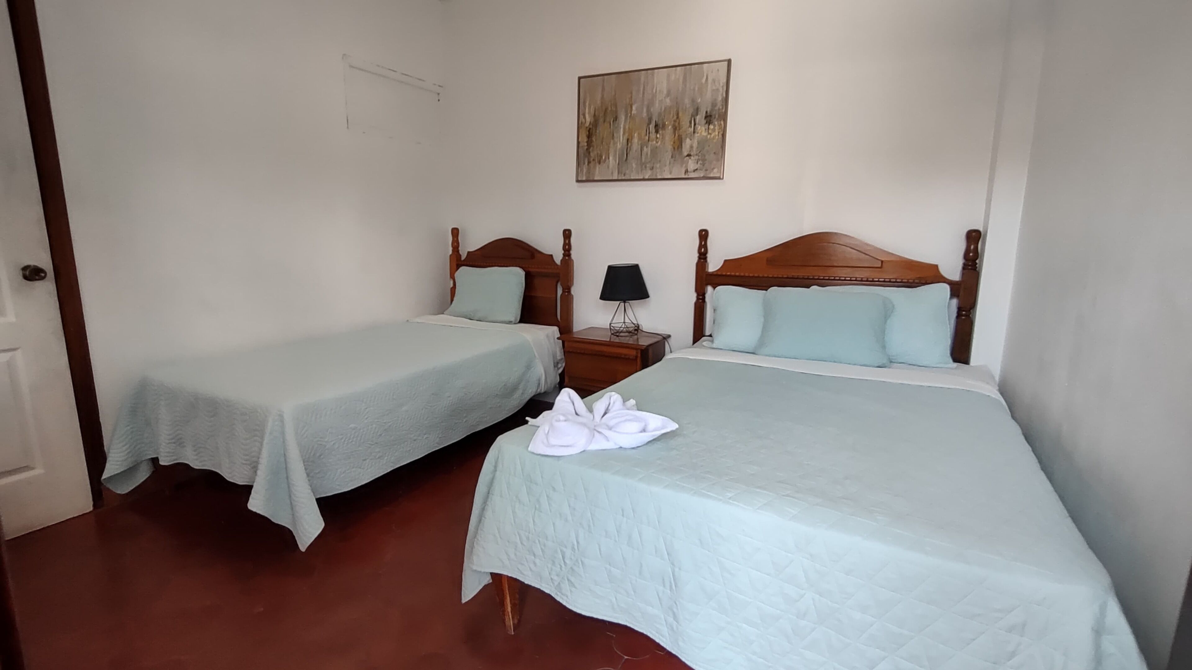 Una habitación de hotel con dos camas individuales, un cabecero de madera, una mesita de noche con lámpara y un cuadro en la pared.