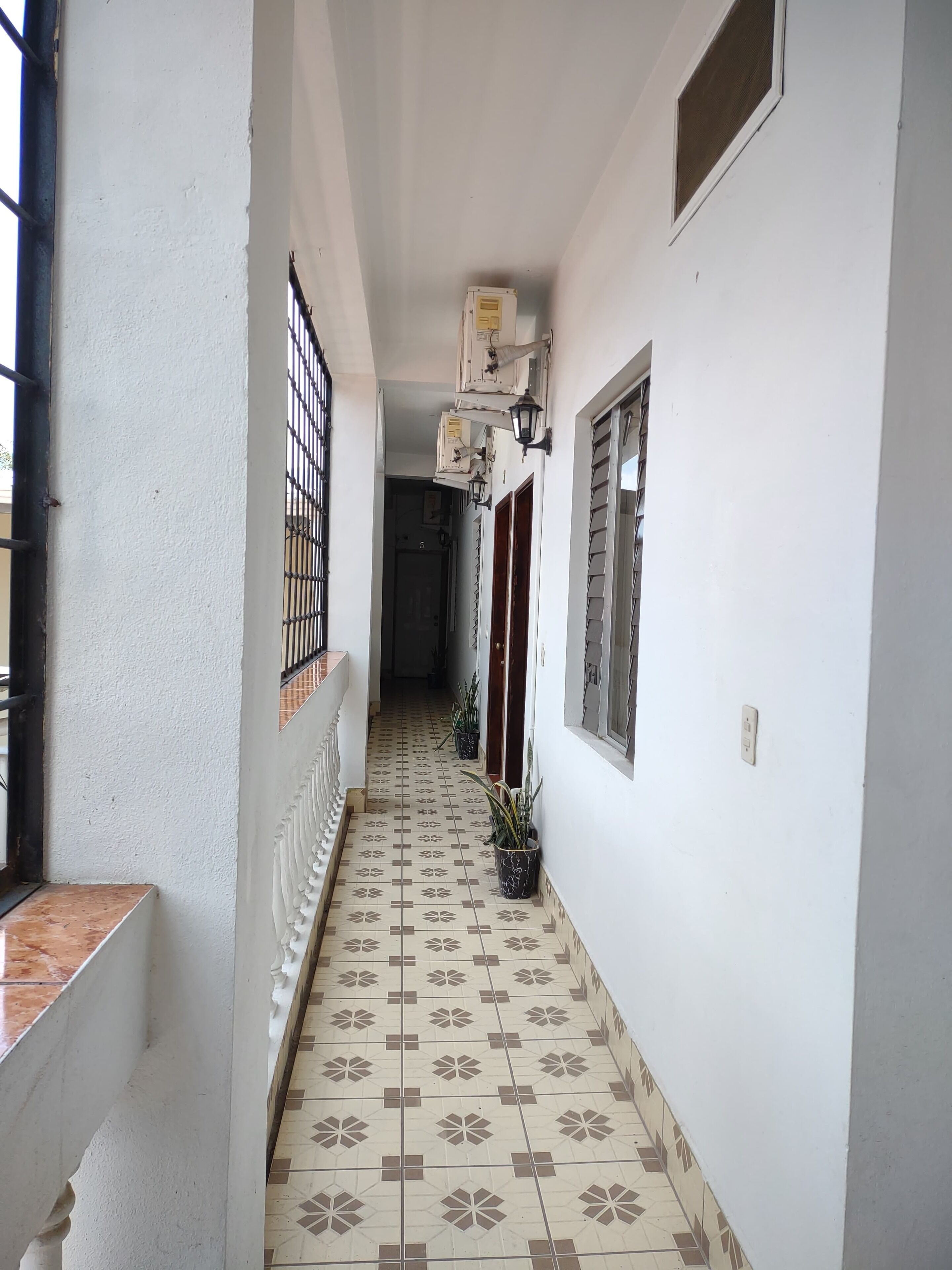 Un couloir étroit, dont le sol est recouvert de carreaux décoratifs, avec une grille métallique sur la gauche et un pot de fleurs sur la droite.