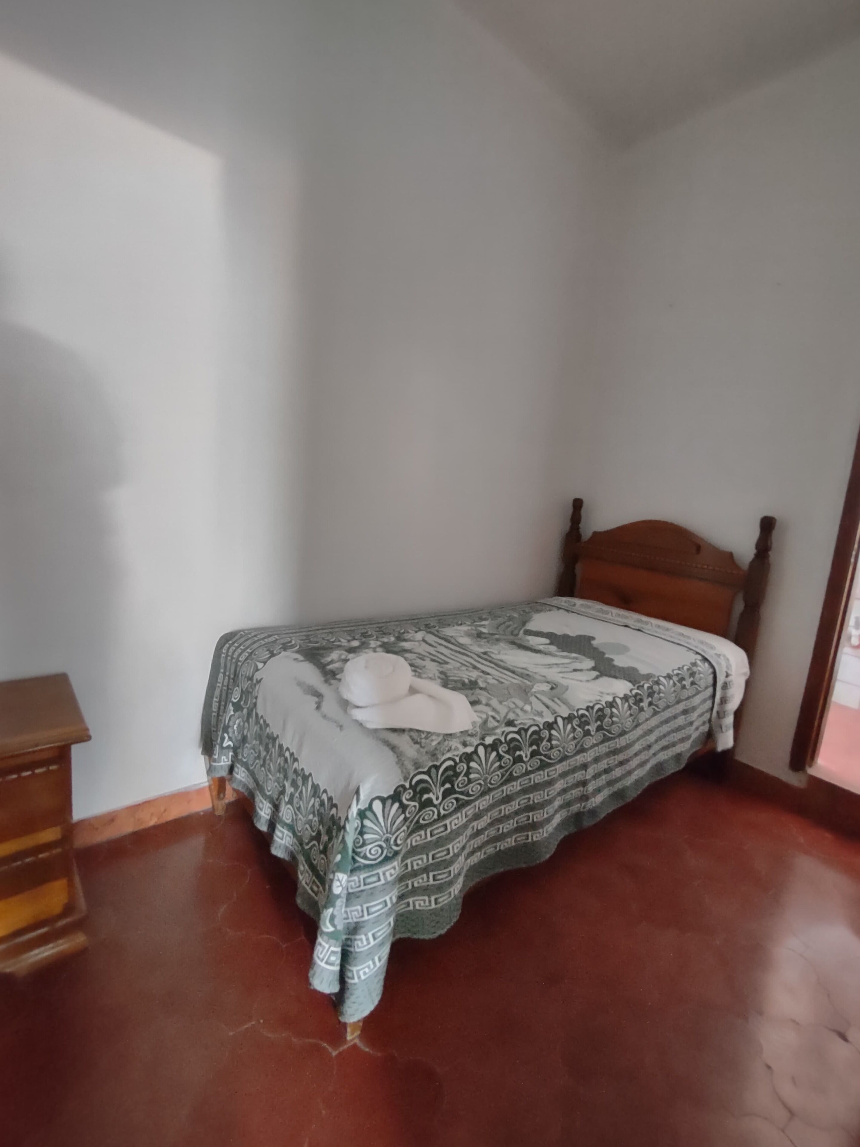 A single bed with a patterned bedspread and a wooden headboard in a room with plain walls and a tiled floor.