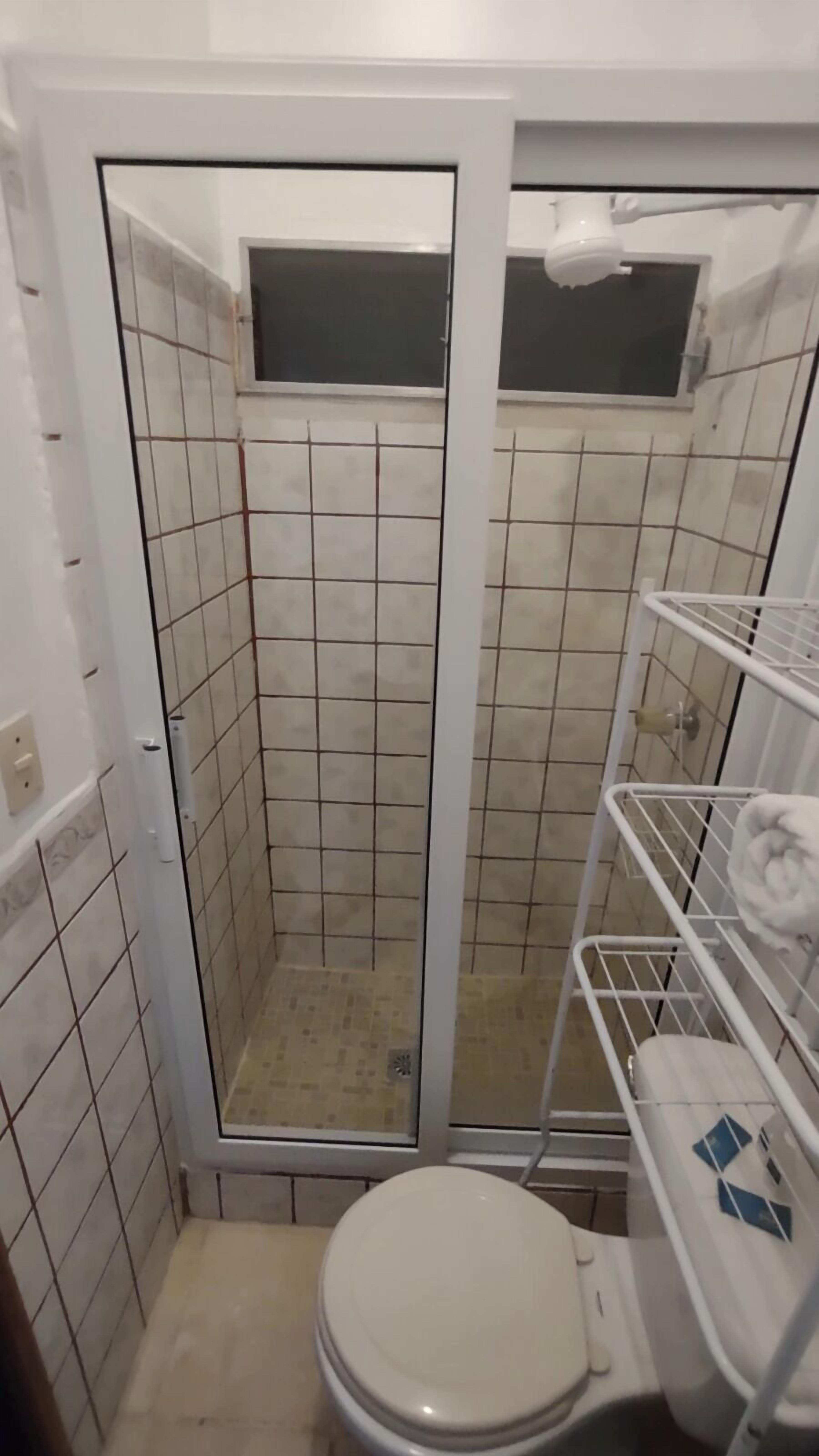 A small, tiled bathroom with a toilet, a shower enclosure, and a shelf with towels.