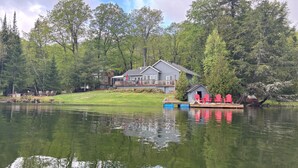 Una cabaña junto al lago con un muelle y sillas rojas, rodeada de un bosque frondoso.