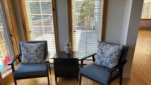 Una habitación con dos sillones azules con cojines estampados, una mesita con un florero y vistas a través de amplias ventanas con persianas.