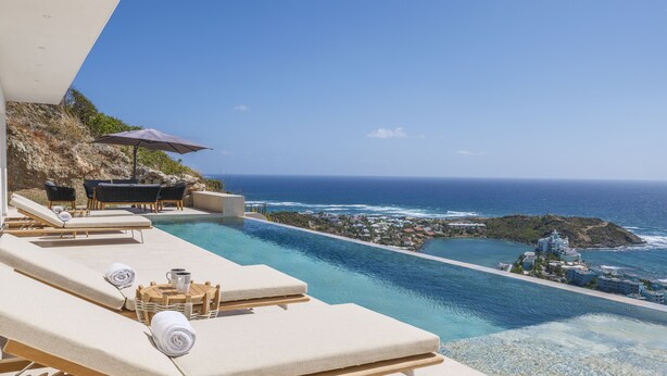 Zona de piscina con sillones reclinables, una mesa y una sombrilla, con vistas a la costa y un cielo despejado.