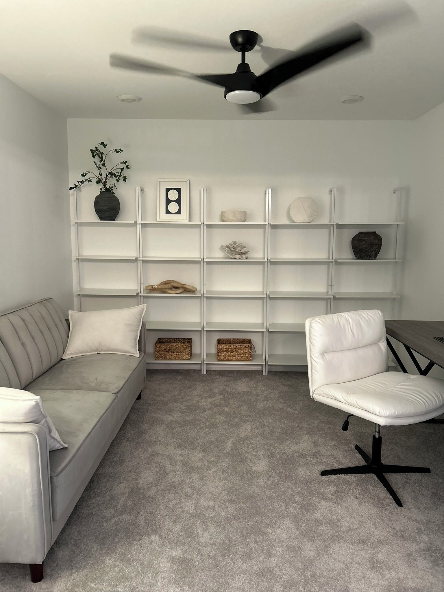 A modern living room with a grey sofa, a white armchair, a white bookshelf, and a ceiling fan.