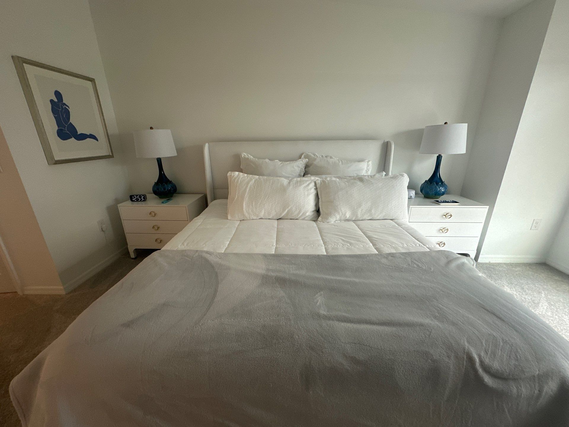 A neatly made bed with white bedding and pillows, two bedside tables with lamps, a wall clock, and a framed picture on the left wall.