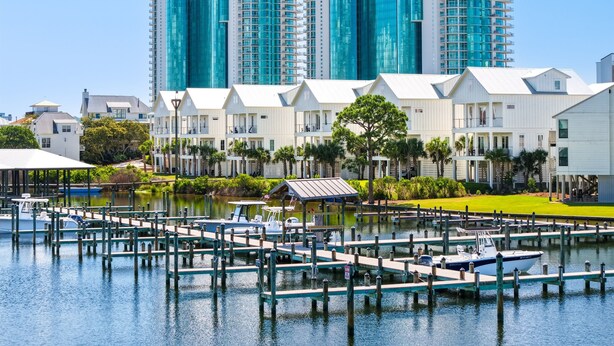 A marina with docked boats, a white building with a flat roof, and a tall blue glass building in the background.