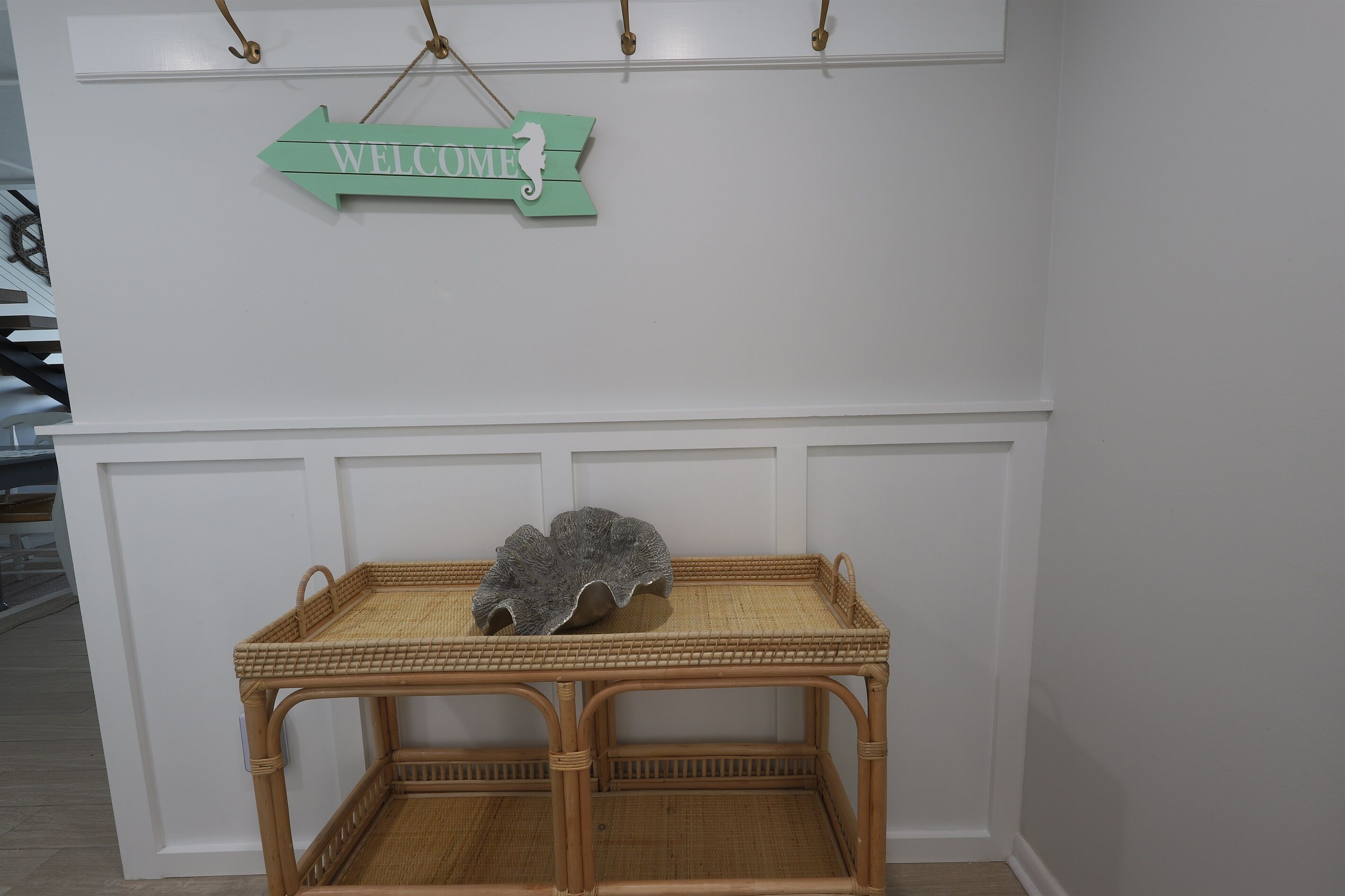A wicker table with a decorative shell centerpiece, a 'WELCOME' sign above, and a white wall with paneling.